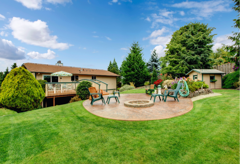 A landscaped backyard with a spacious green lawn, a circular brick patio with outdoor chairs and a fire pit, and a residential deck surrounded by mature trees and colorful garden plants.